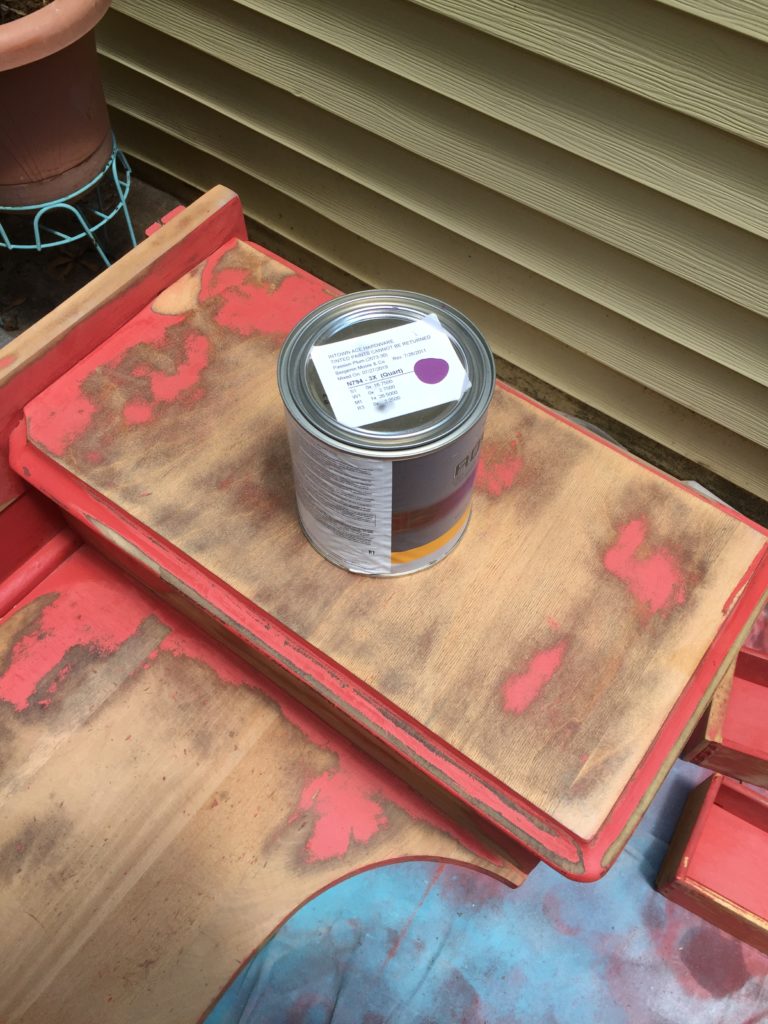 An unopened can of purple paint on top of a partially sanded-down wooden vanity with patches of red paint still remaining.