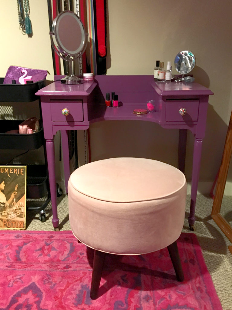 Purple vanity and pale pink stool on a bright pink rug, with makeup supplies.