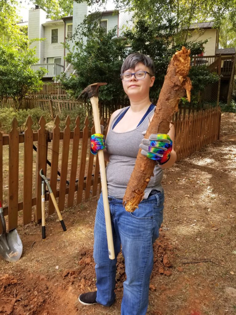 Indigo stands proudly, holding the mattock in one hand and a tree root the size of their arm in the other.